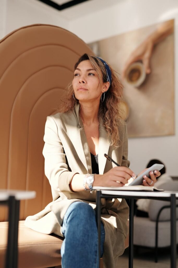 Confident woman in blazer taking notes with pen and smartphone in cafe.