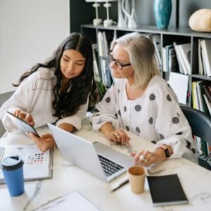 Two professional women discussing work on a laptop and tablet in a contemporary office setting.