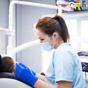 A female dentist wearing gloves and a mask attends to a patient in an indoor clinic setting.